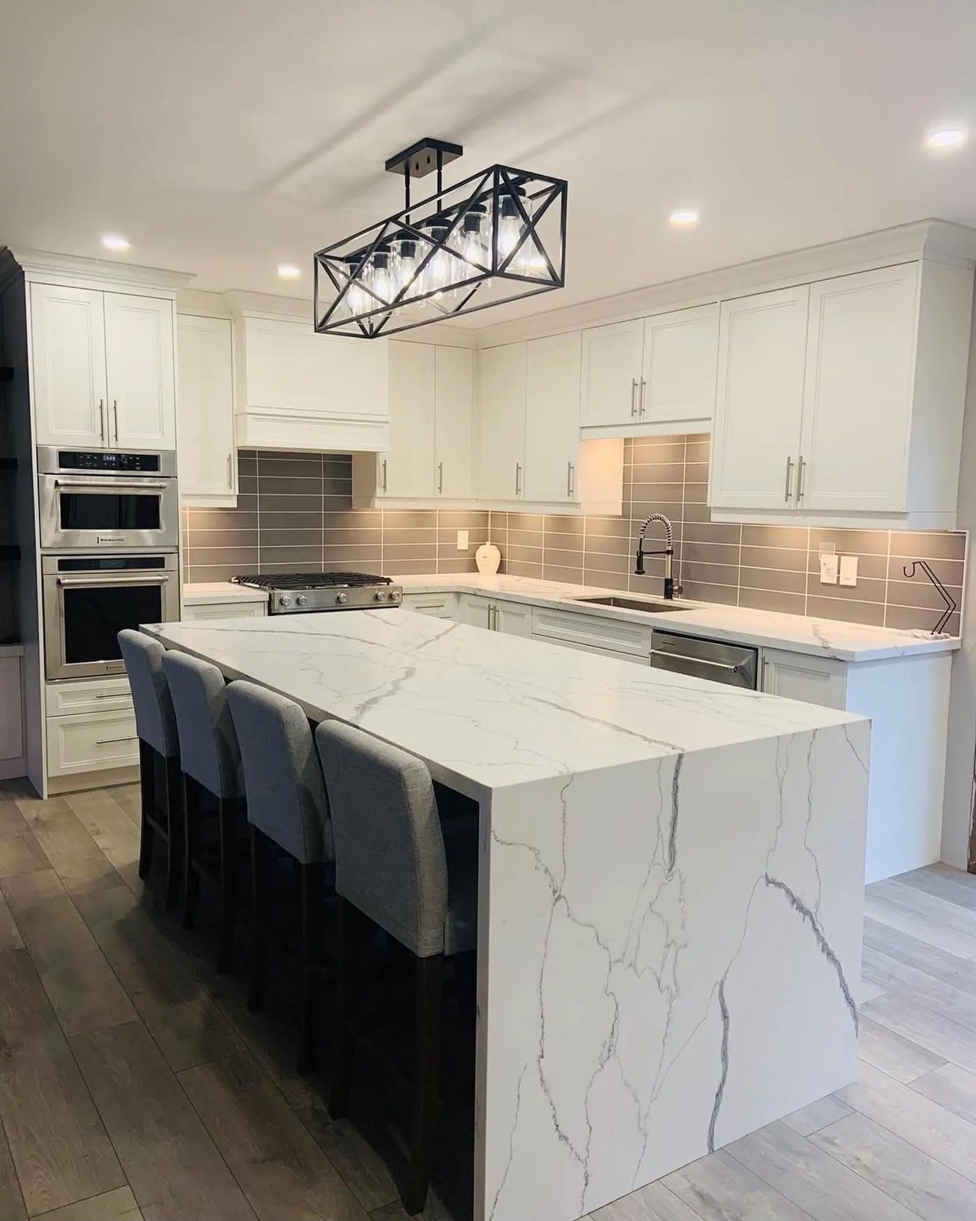 Modern white kitchen with a large marble waterfall island, grey bar stools, and geometric chandelier.