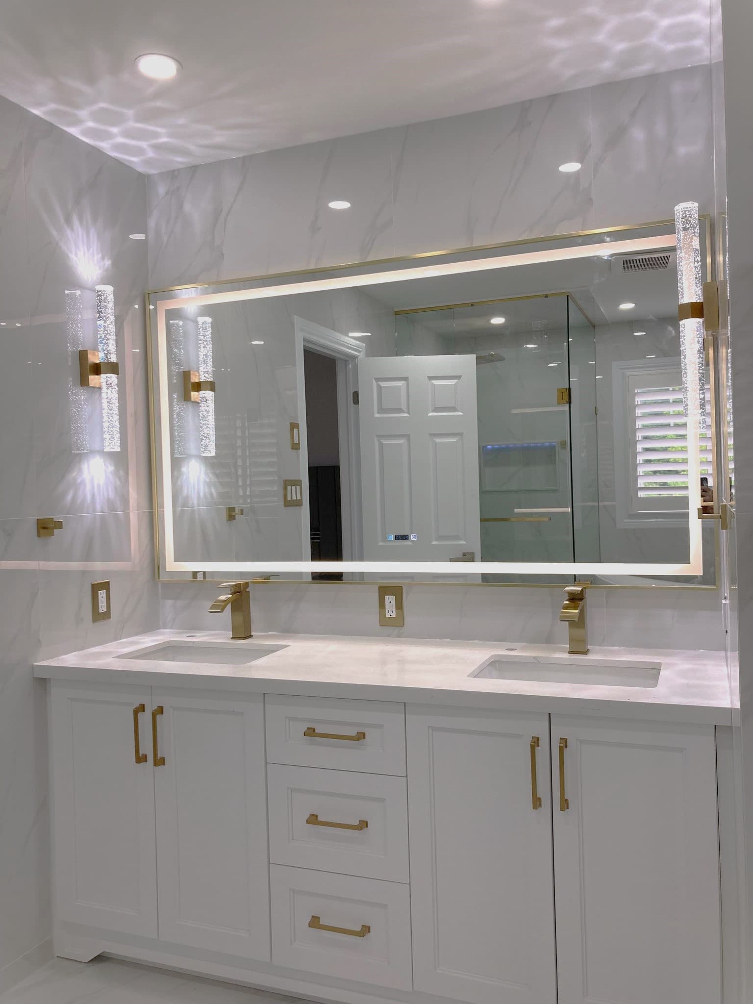 Modern white double vanity featuring gold hardware, marble walls, and a large backlit mirror.