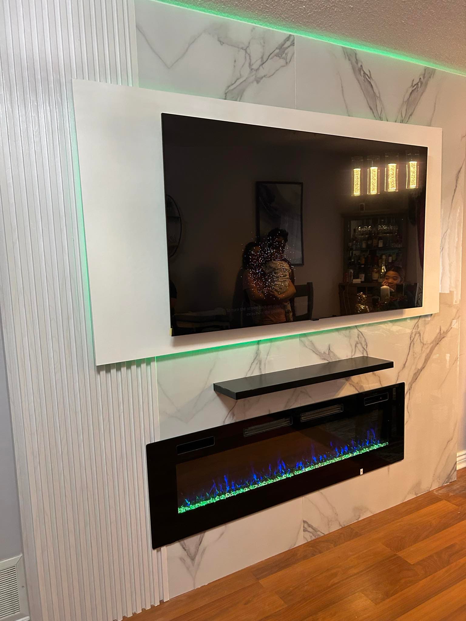Wall-mounted TV on marble panels with green LED backlighting above a modern electric fireplace.