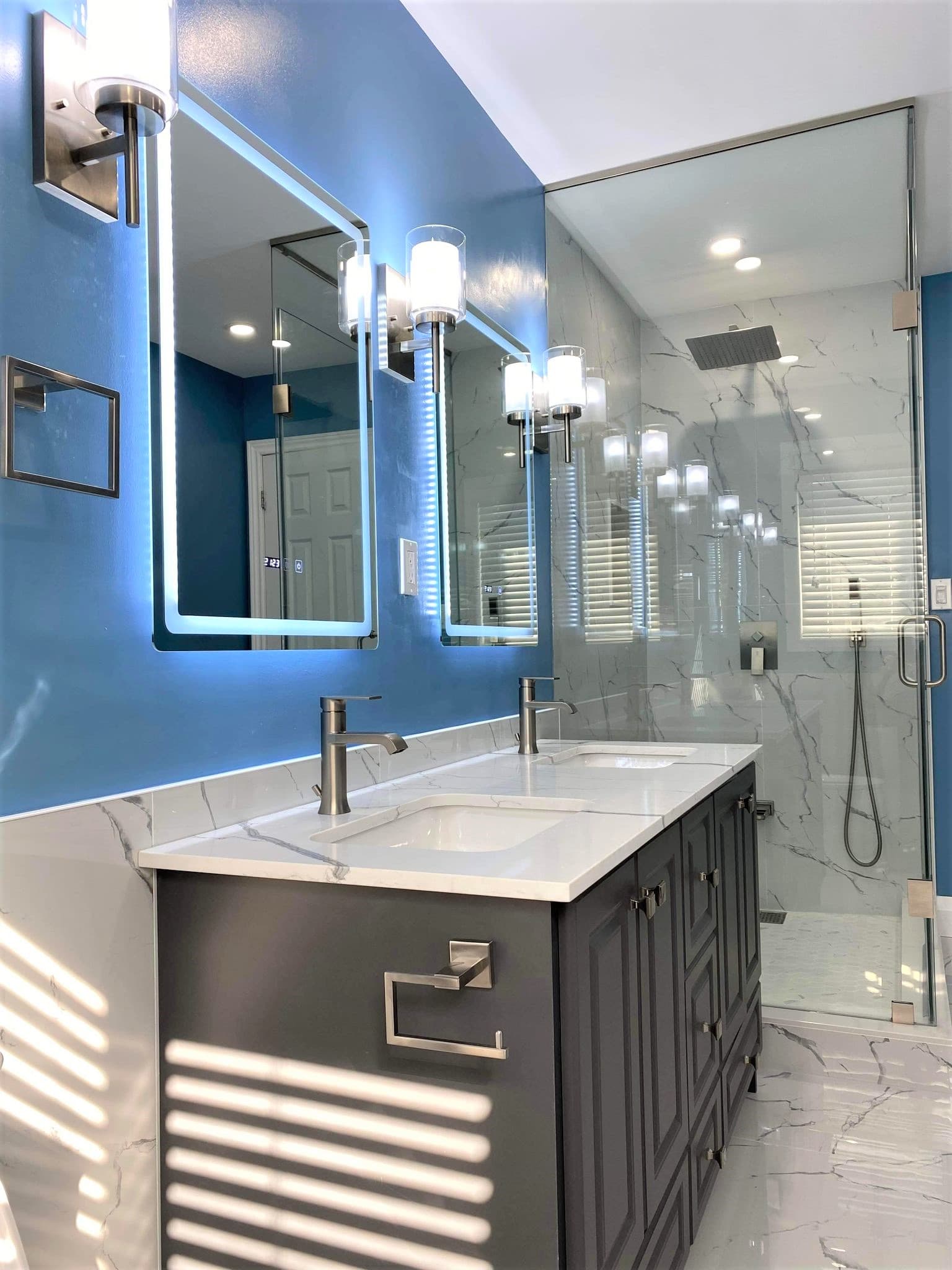 Modern bathroom with blue walls, double vanity, backlit mirrors, and glass-enclosed marble shower.