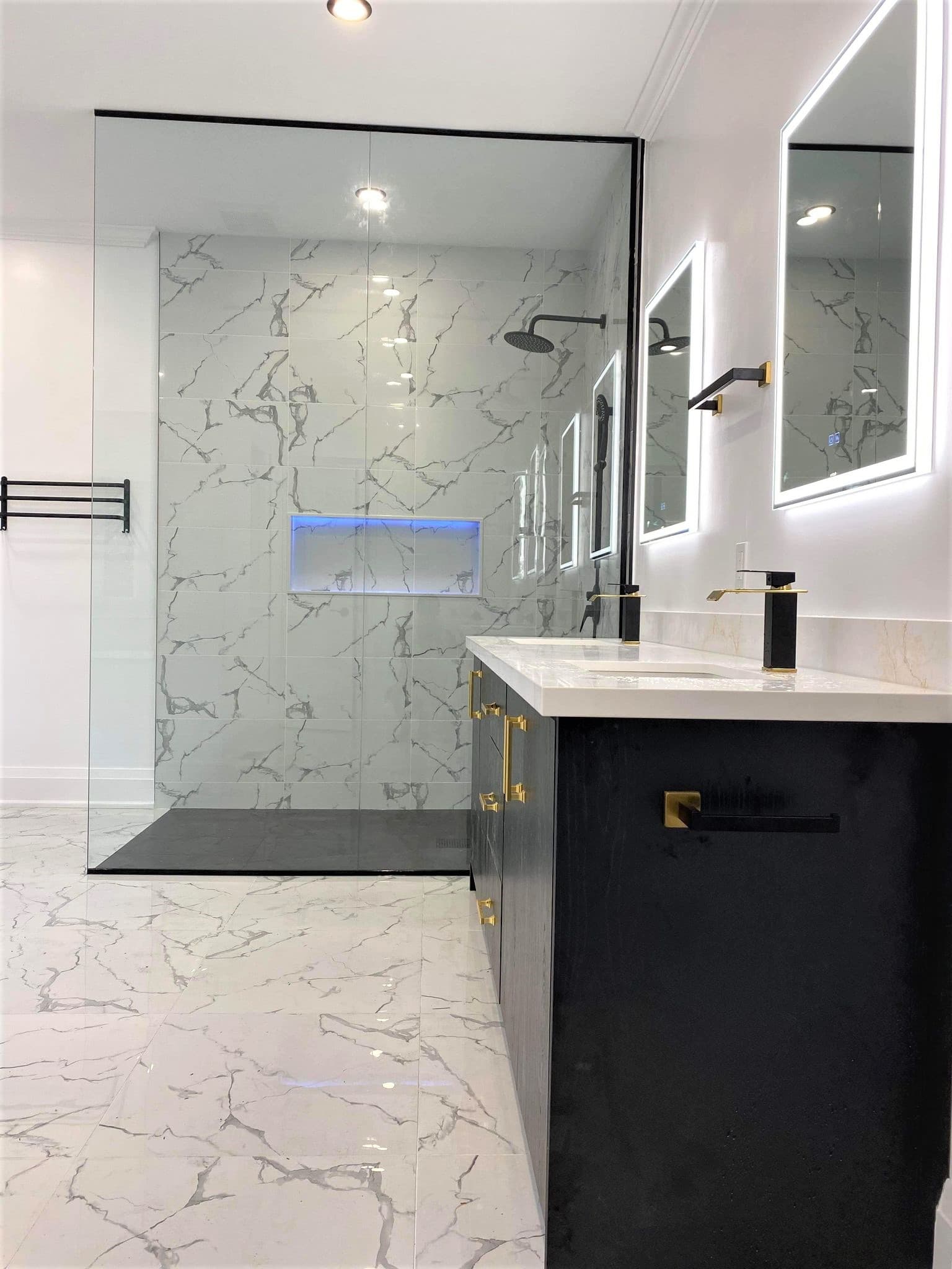 Modern bathroom featuring marble tiles, glass shower, black vanity, gold fixtures, and backlit mirrors.