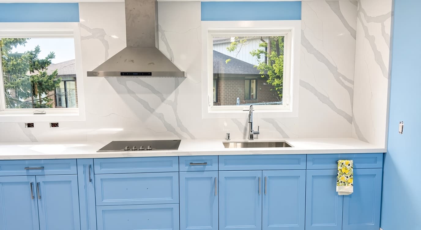 Modern kitchen featuring light blue cabinets, white marble backsplash, stainless steel hood, and sink.